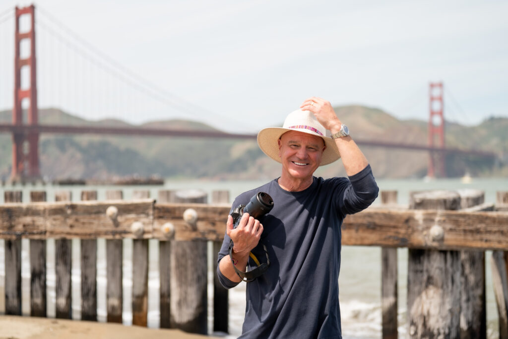 A photographer holding a camera and smiling next to the Golden Gate Bridge