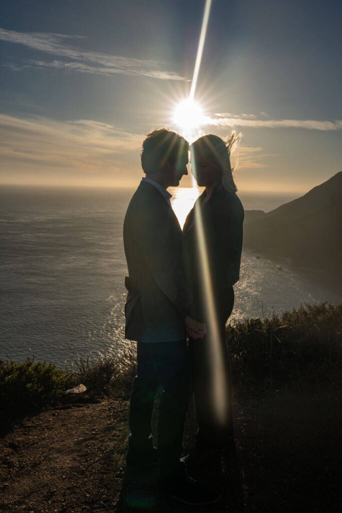 Couple embracing at sunset with dramatic light flaring over the Pacific Ocean near San Francisco