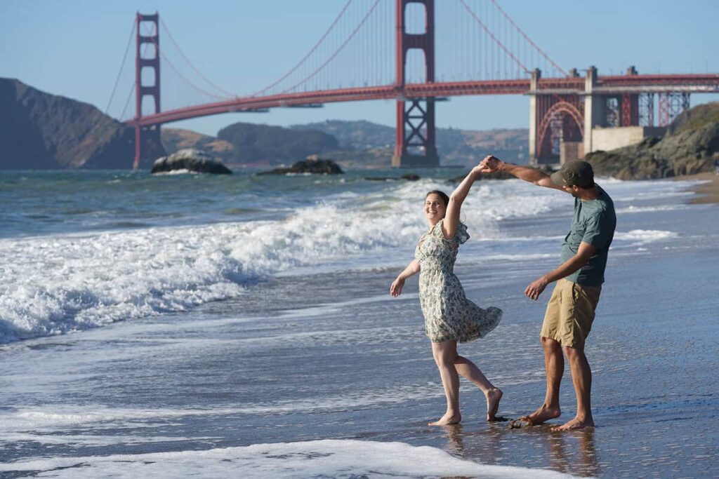 Couple running hand-in-hand along Baker Beach with the Golden Gate Bridge in the background at sunset