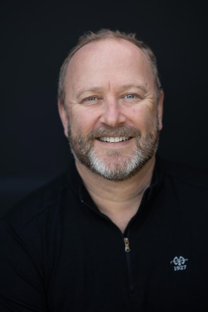 Male executive smiling in a professional portrait with a black shirt against a dark background