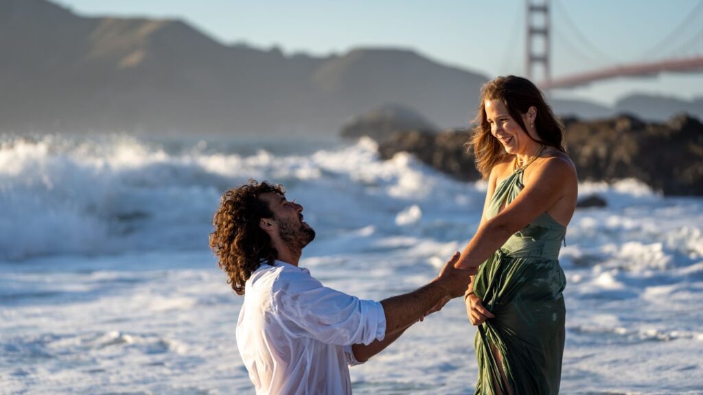Man proposes to woman at Baker Beach with the Golden Gate Bridge in the background at sunset.