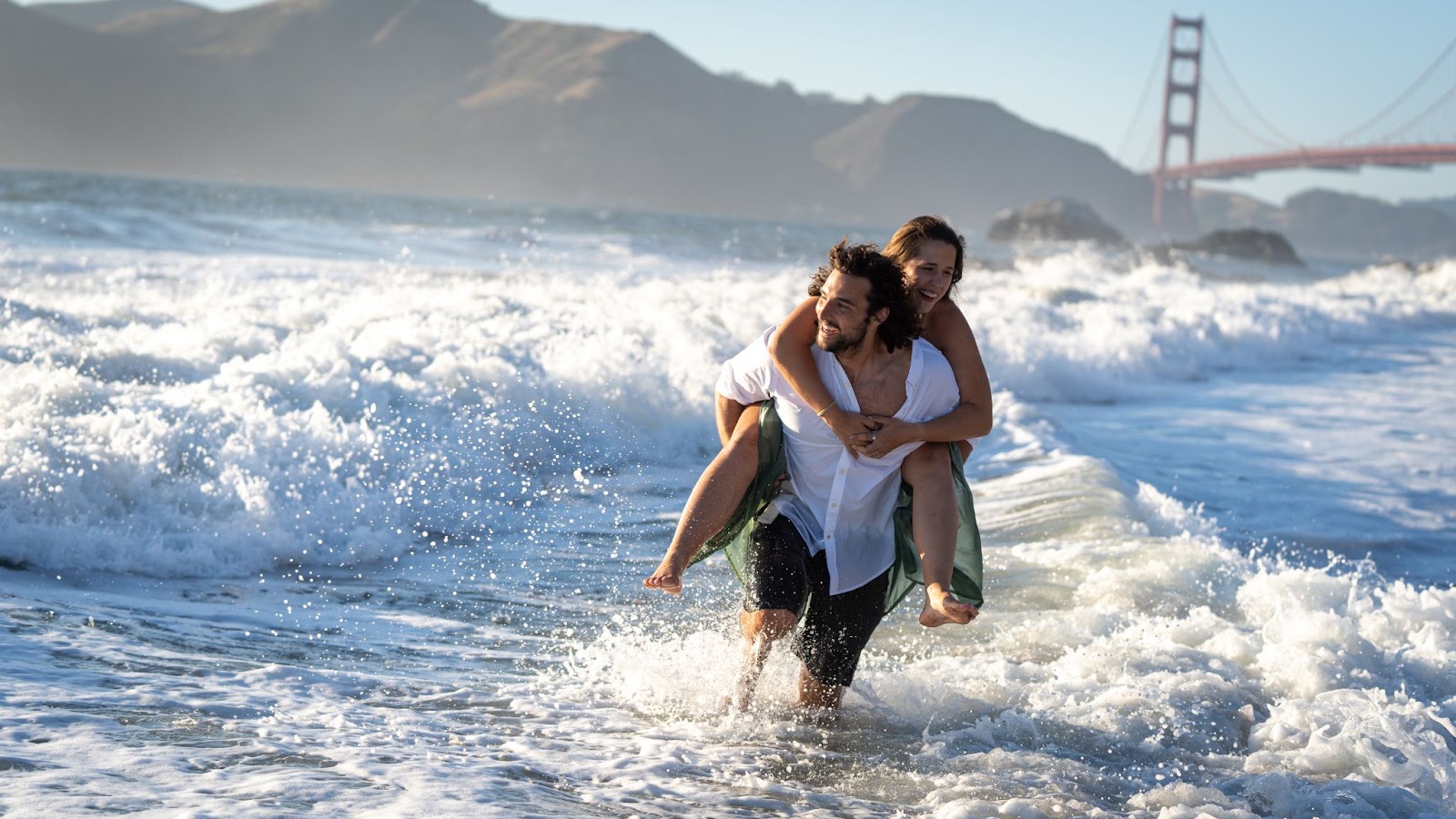 Couple walking through ocean waves at Baker Beach with the Golden Gate Bridge in the background