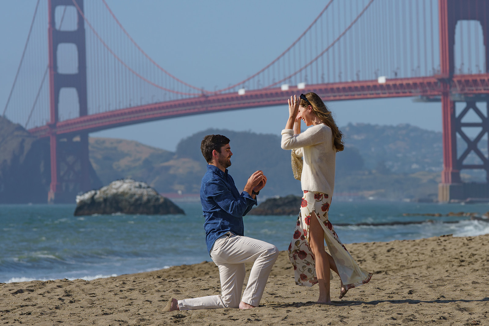 Man on one knee at the beach proposing to a woman in shock from the surprise with the golden gate bridge as a backdrop.