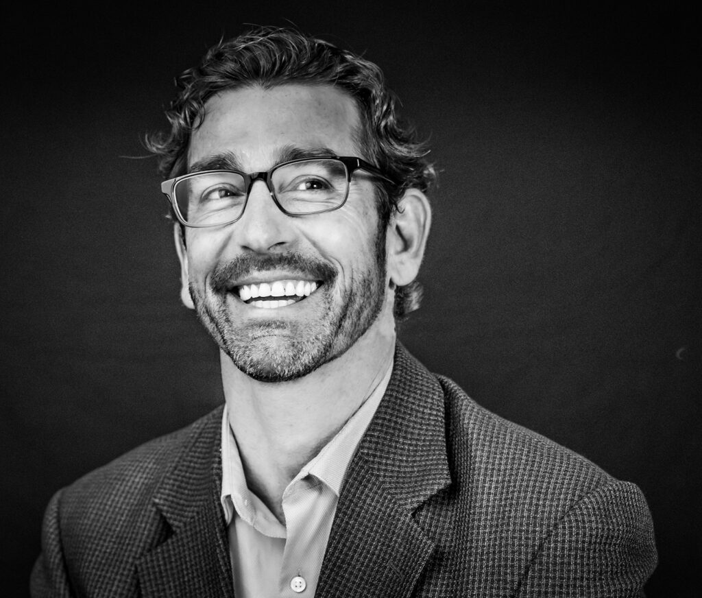 Smiling man in glasses with a beard, wearing a blazer — corporate headshot on dark background