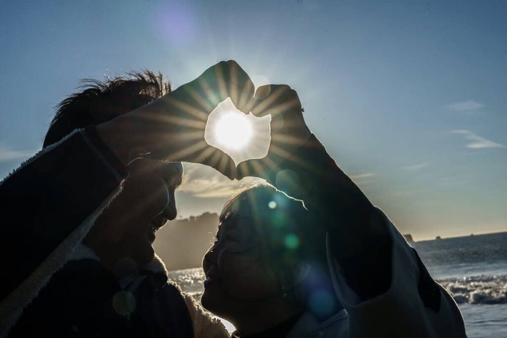 Silhouetted couple forming a heart shape with their hands in front of the setting sun near the ocean