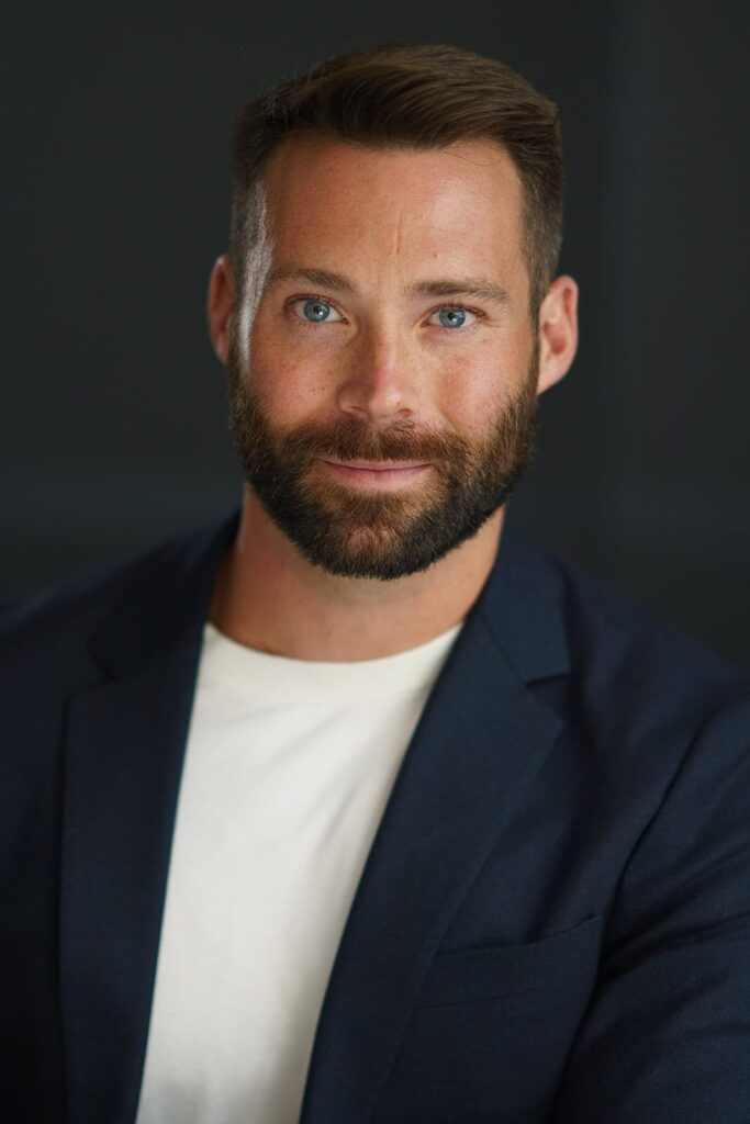 Confident man with trimmed beard in blazer and white t-shirt — studio headshot with dark background

