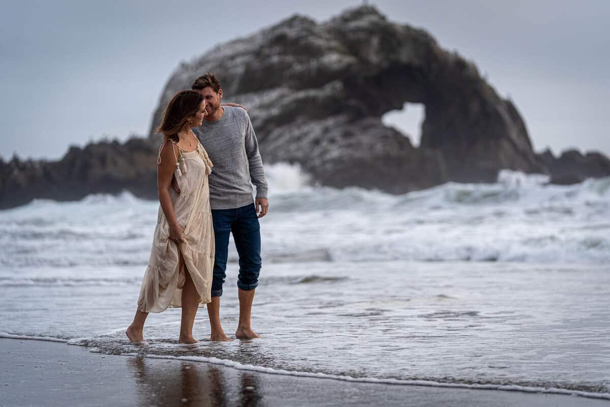 Couple walking along the shoreline at Sutro Baths with waves and rocky cliffs in the background